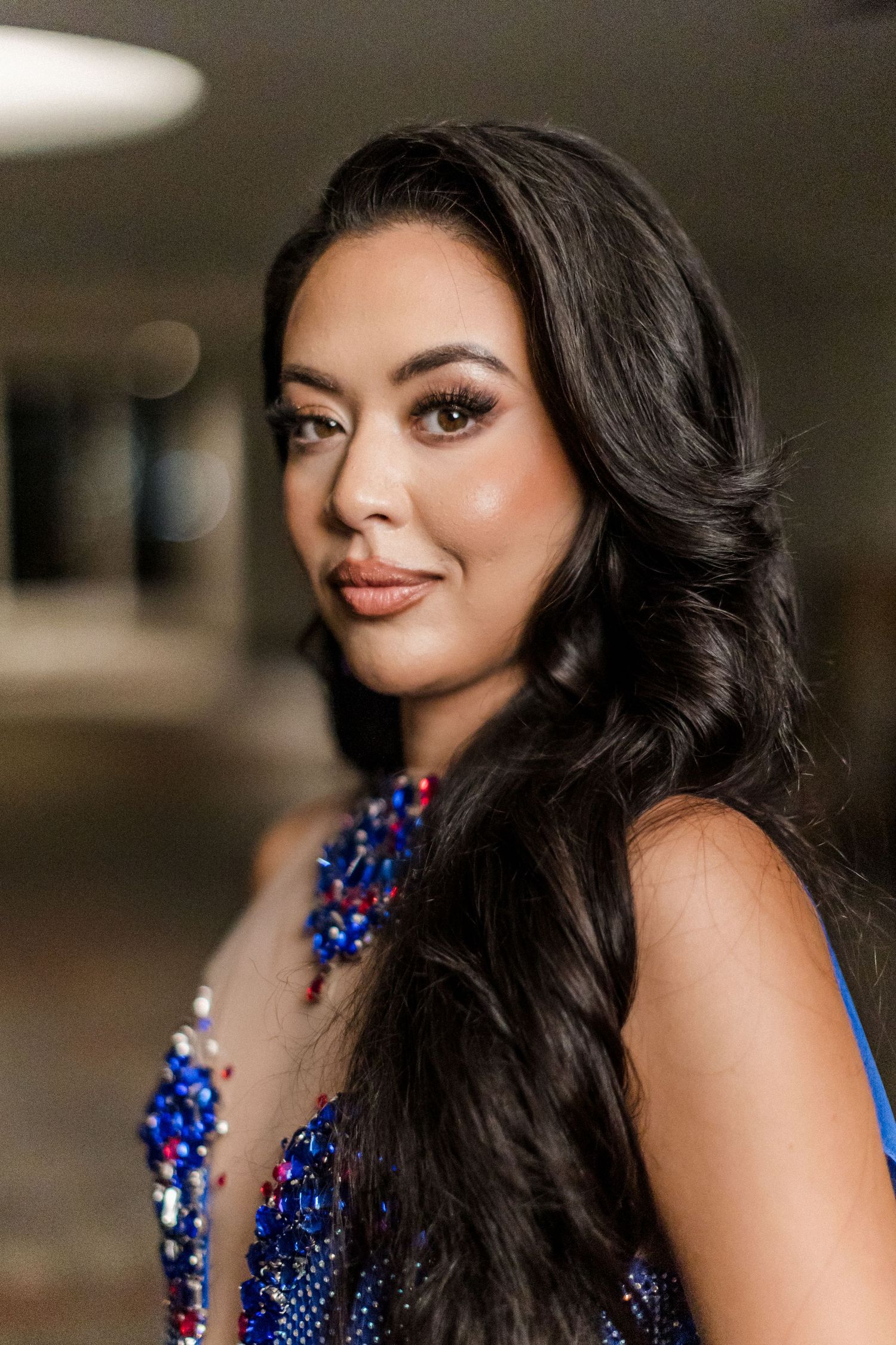 A woman in a sparkling blue gown with long dark wavy hair poses for an elegant portrait.