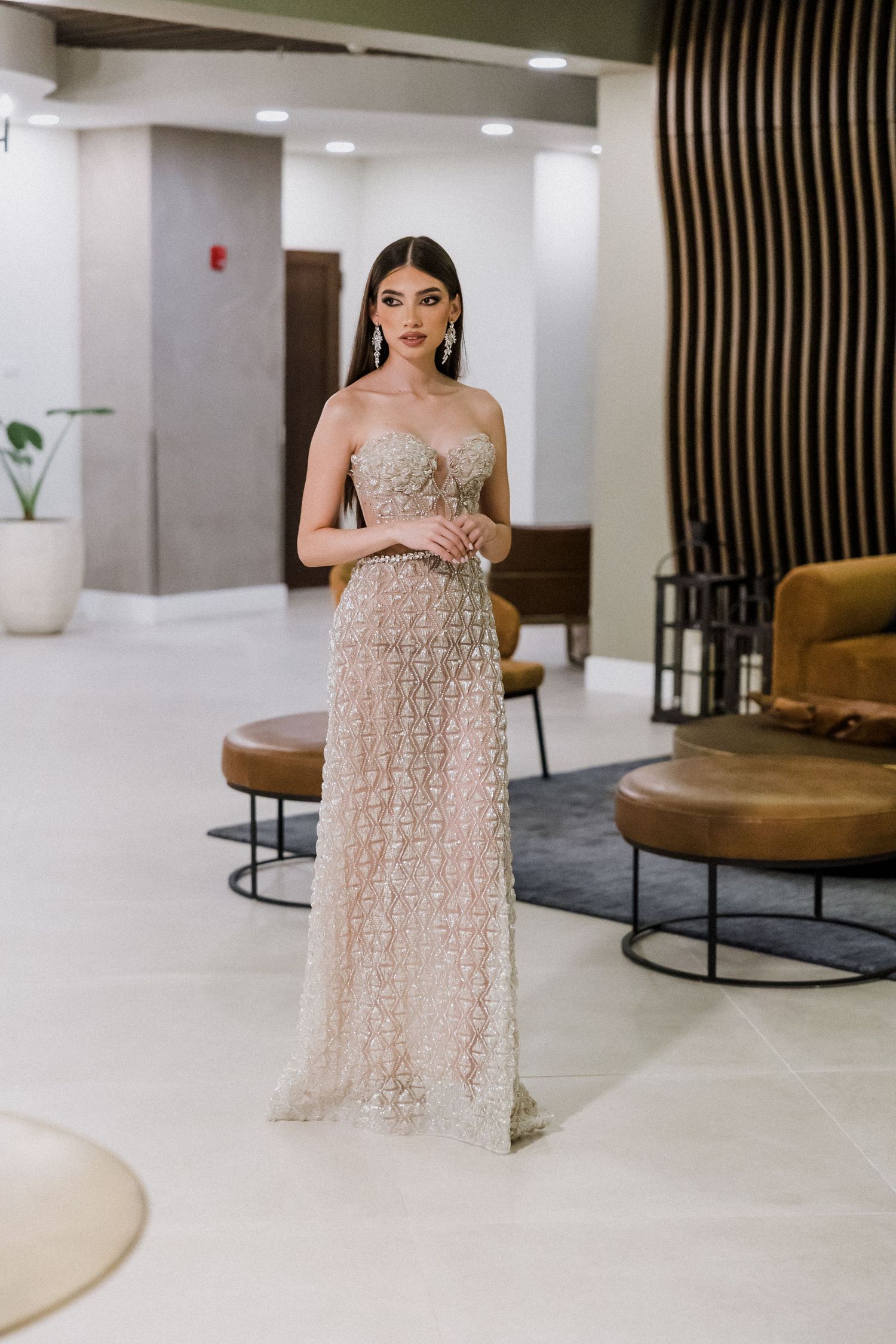 Woman wearing an elegant strapless beige tulle gown stands in a modern lobby with circular brown chairs and white floors.