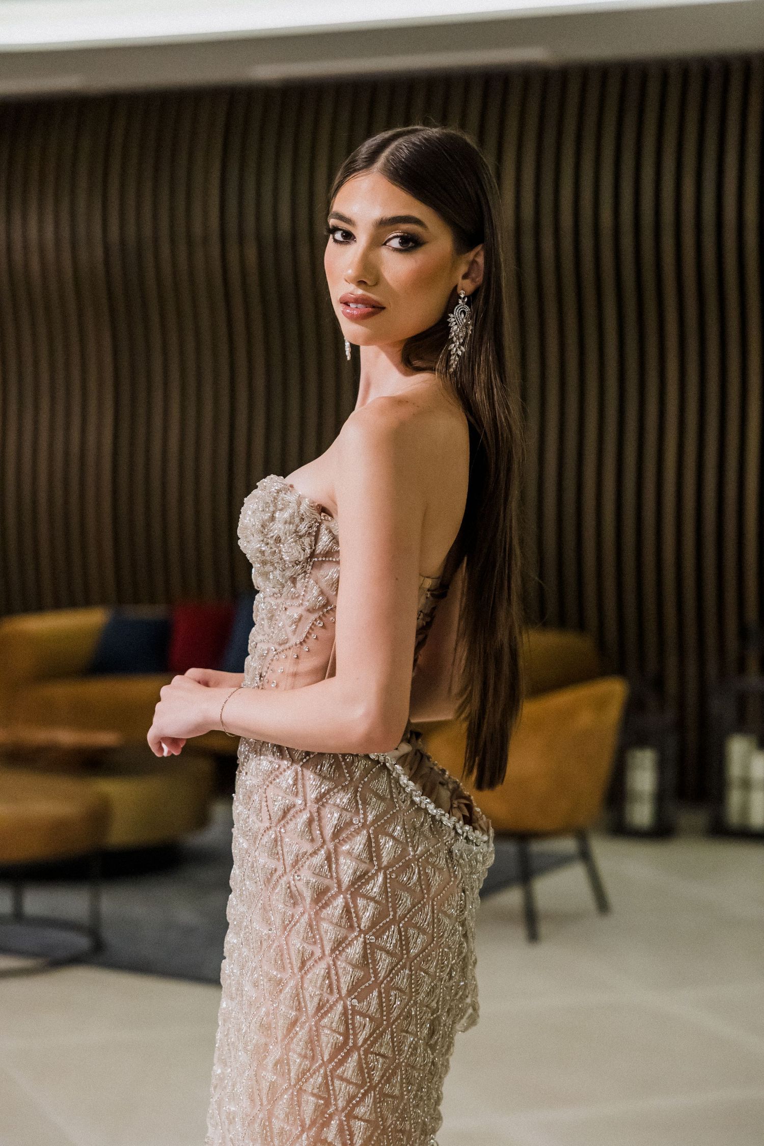 Elegant model poses in a strapless beige beaded evening gown with long straight hair and dangling earrings.