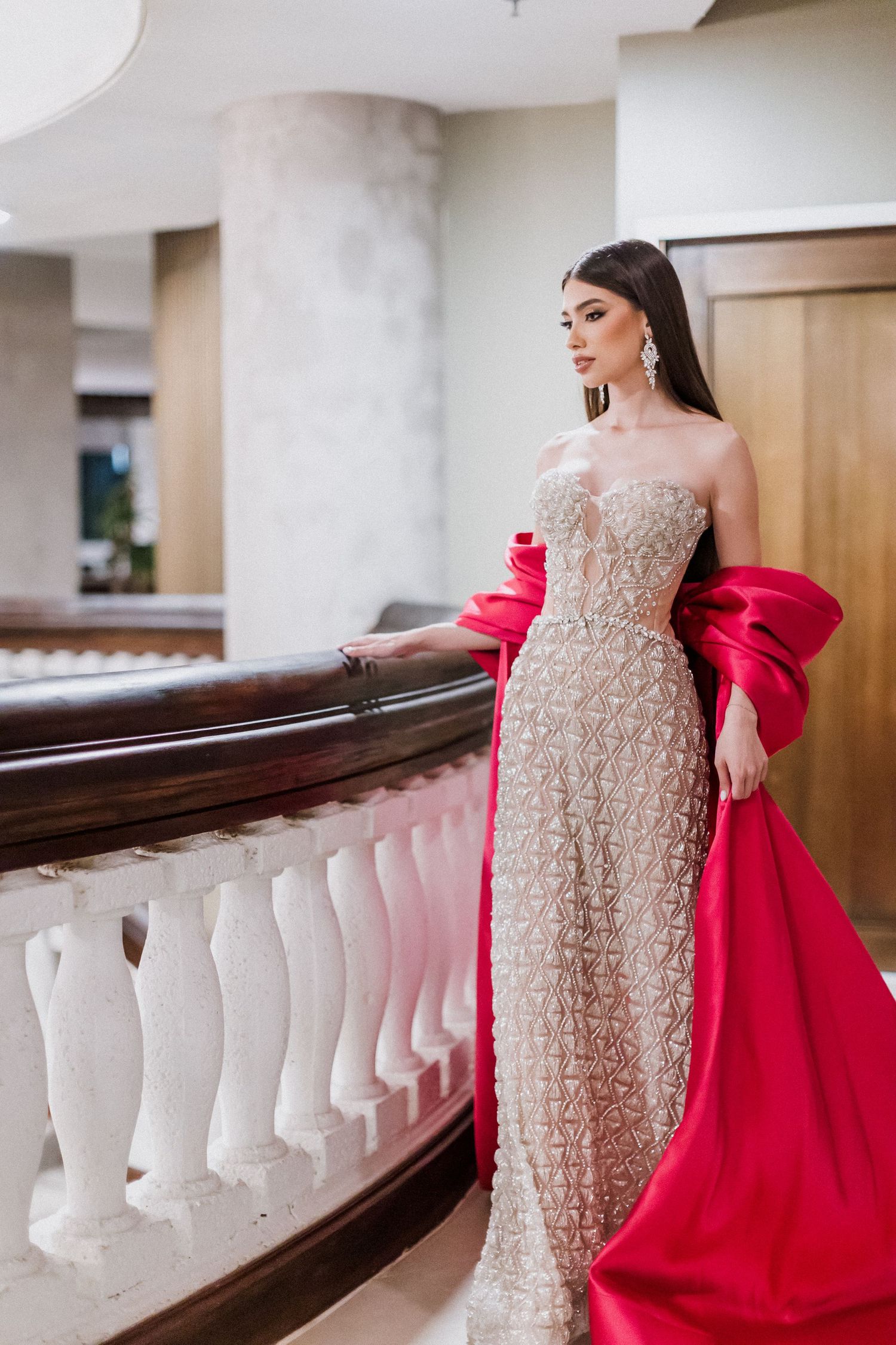 Glamorous evening gown in beige lace with dramatic red cape accent against white balustrade in elegant interior setting.