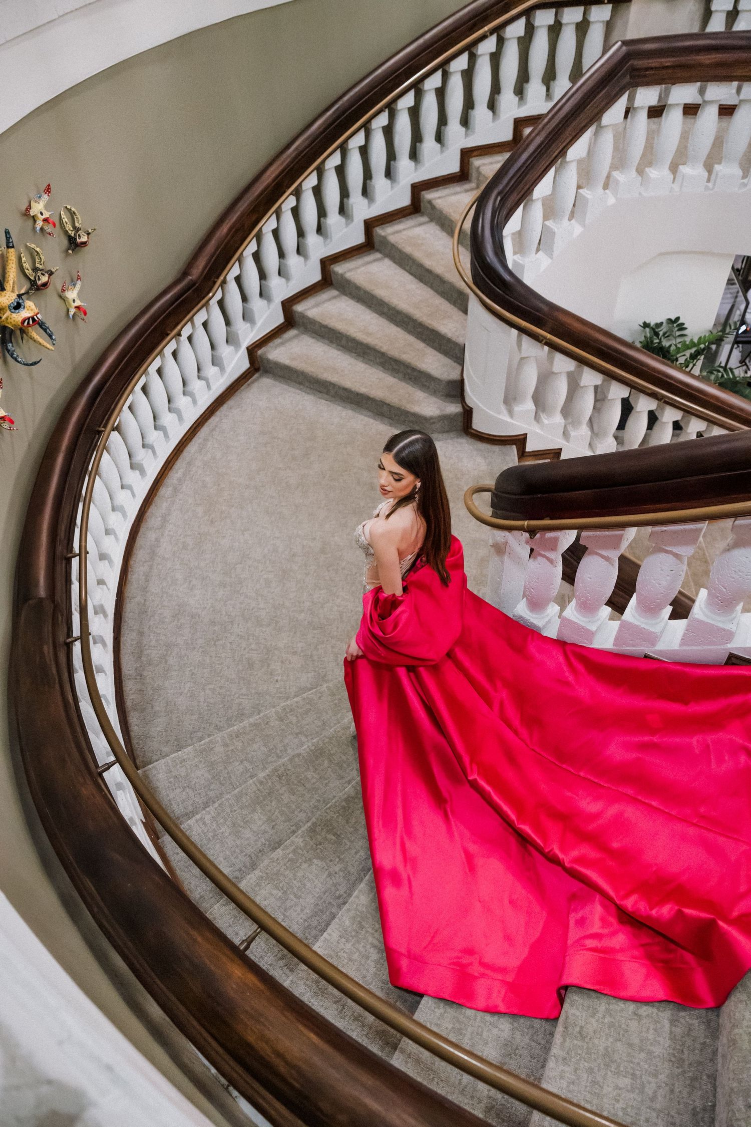 Elegant red ballgown dress flows down a curved grand staircase with white railings and dark wood banisters.