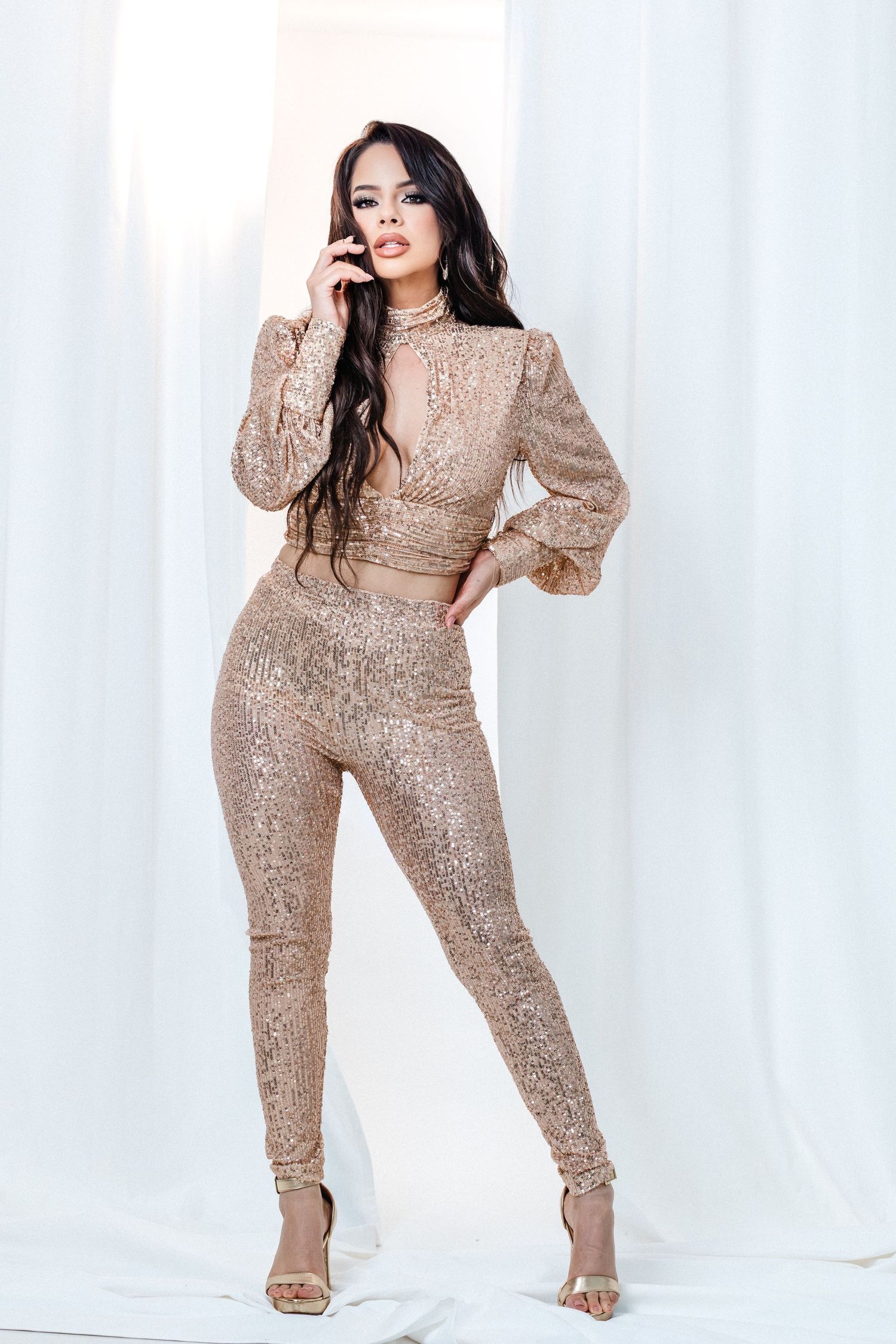Glamorous long sleeve sequined jumpsuit in rose gold with fitted silhouette and high neck design against white backdrop.
