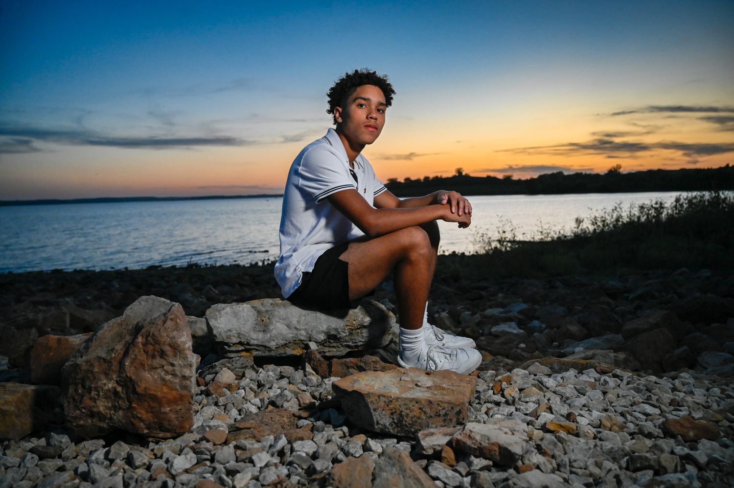 A person sits on coastal rocks watching a vibrant sunset over the water.