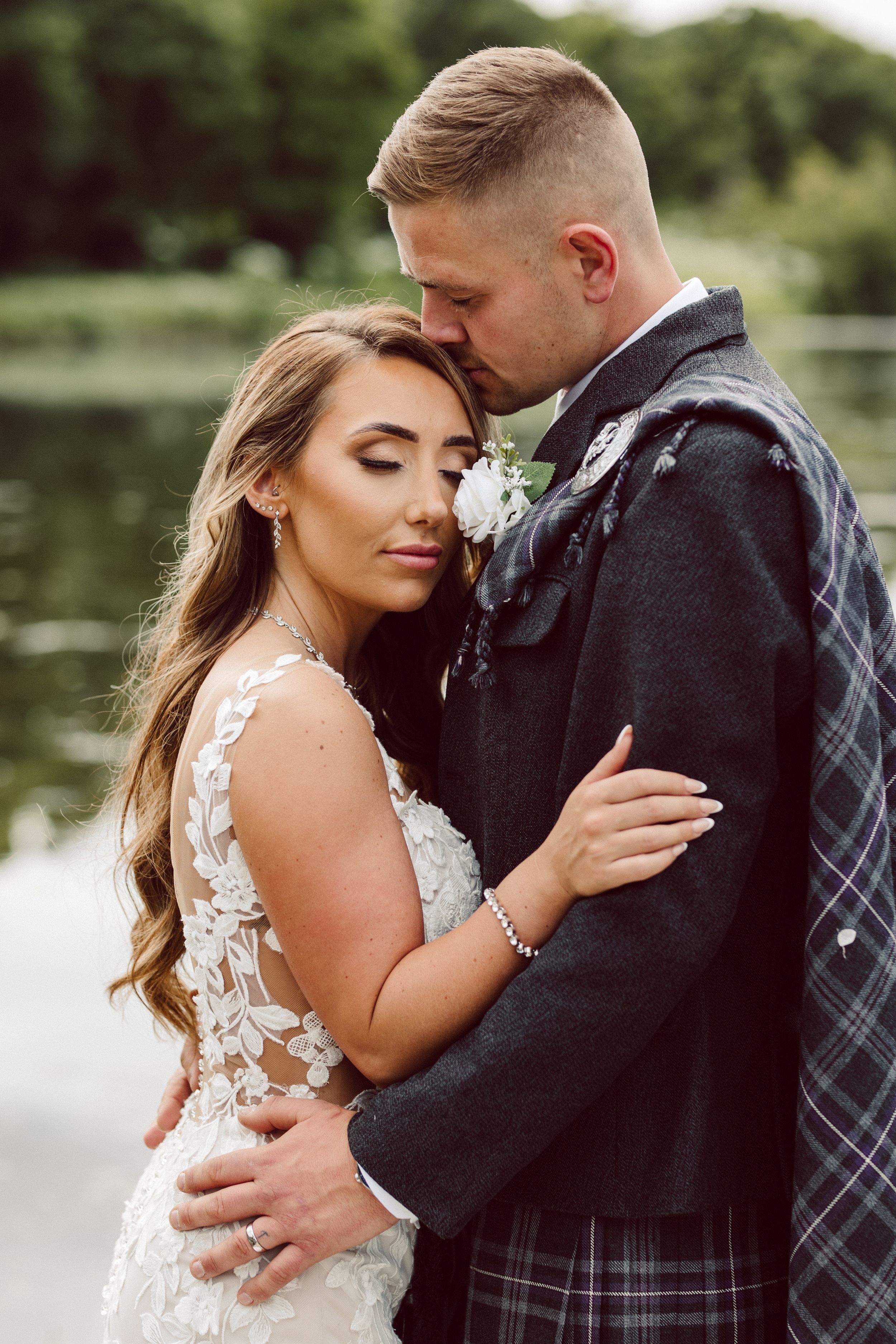 Romantic wedding couple embraces by a serene lake in traditional Scottish attire with lace details.