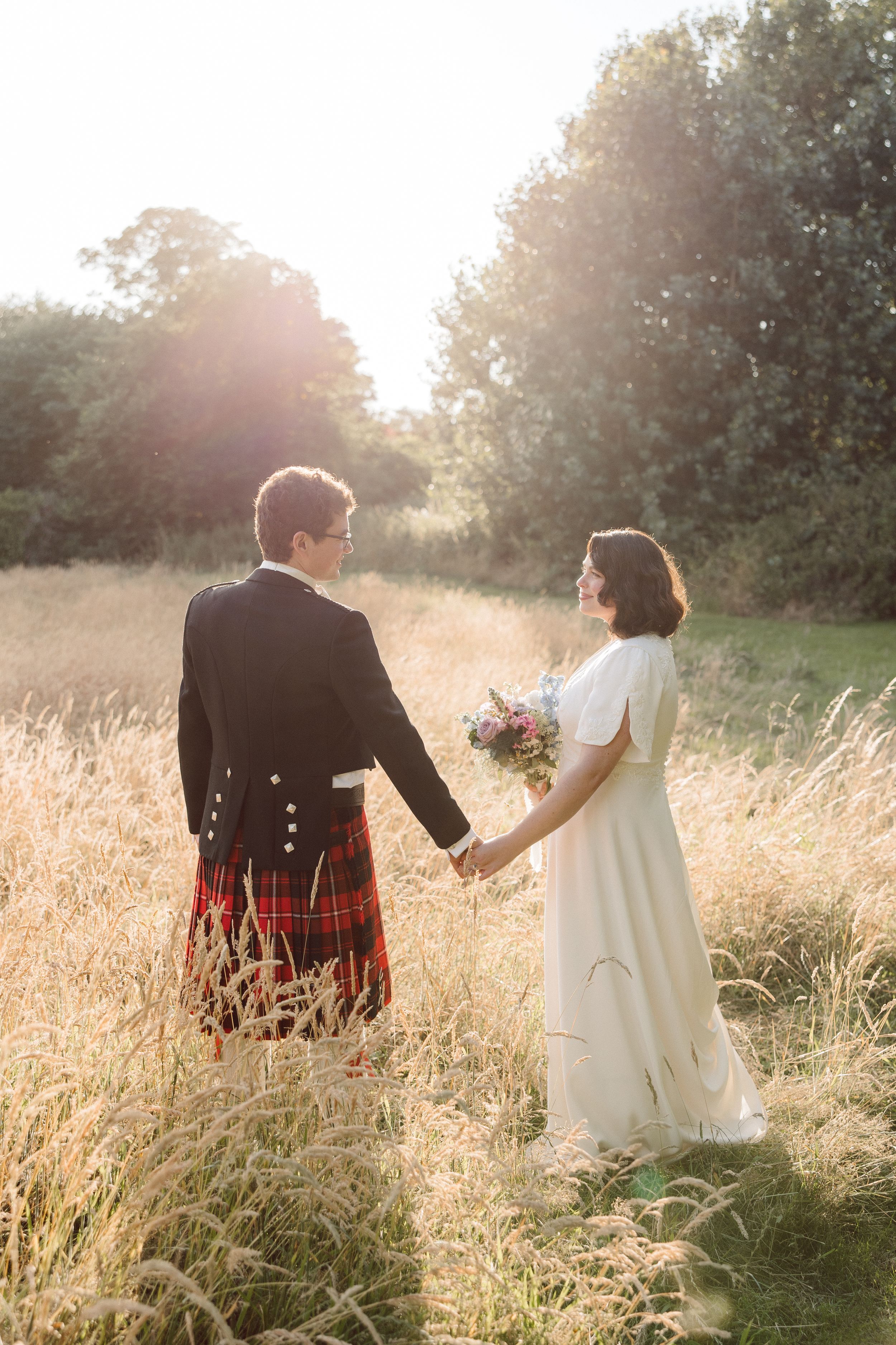 Couple walks through golden wheat field at sunset in romantic wedding attire.