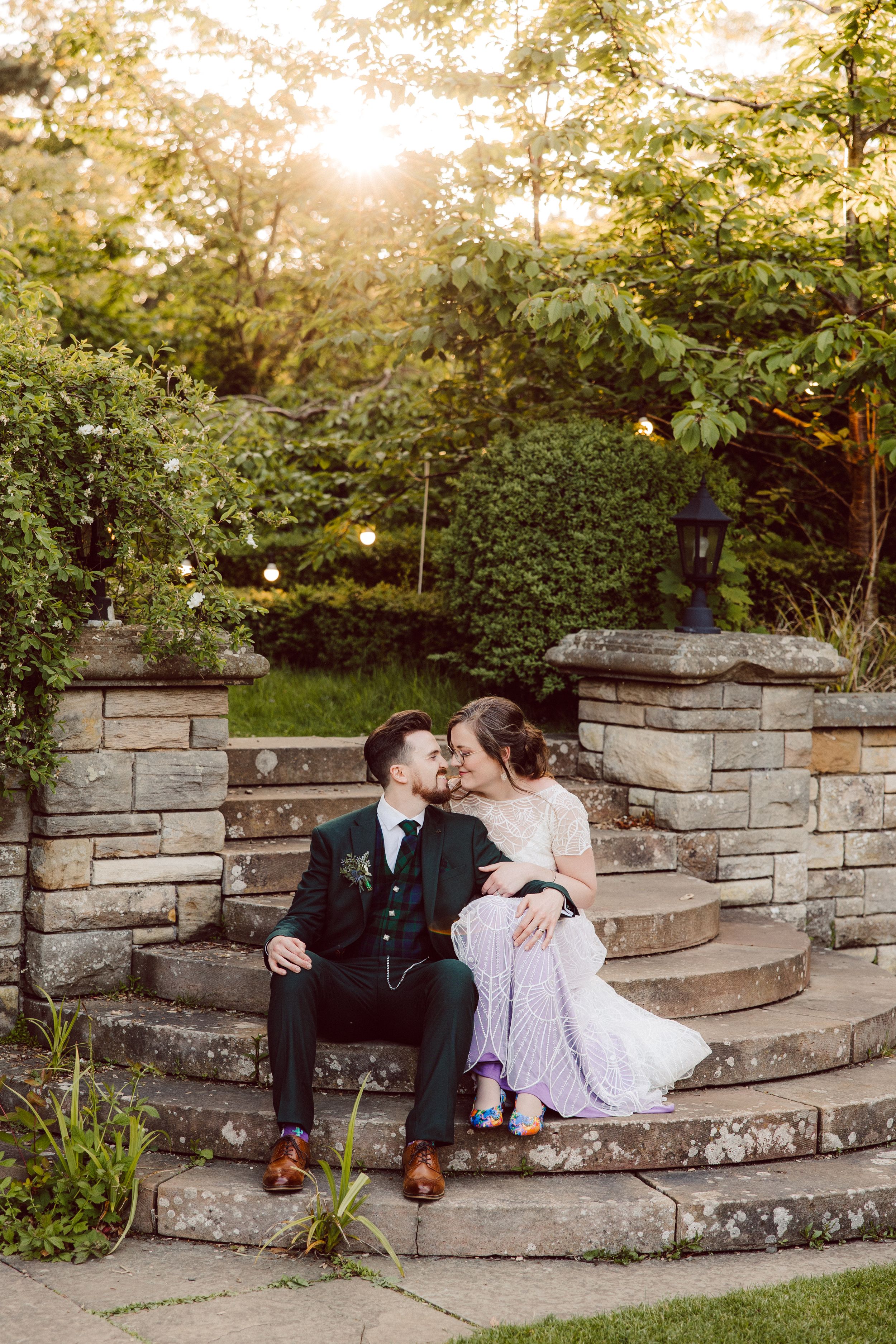 Bride and groom share a romantic kiss on stone garden steps at golden hour, surrounded by lush greenery.