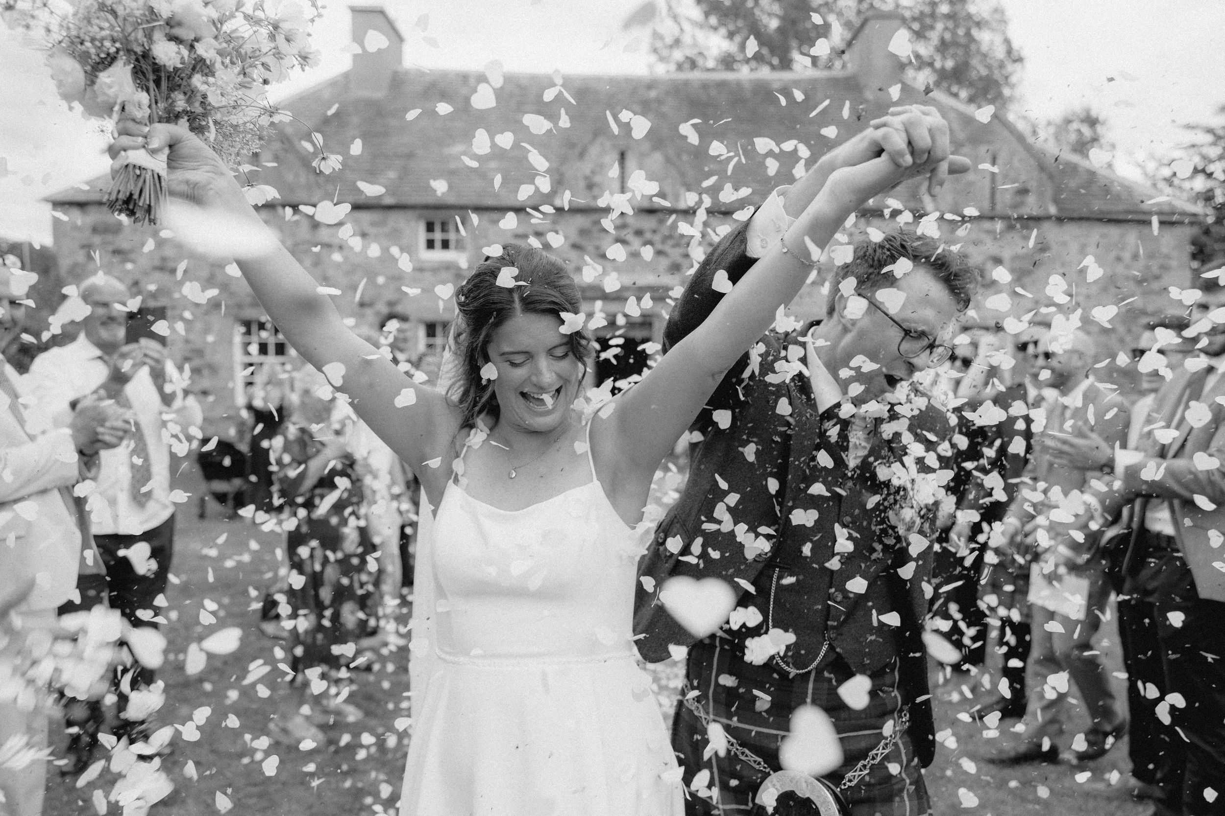 A joyful bride raises her arms in celebration as white confetti falls around her in this black and white photograph.