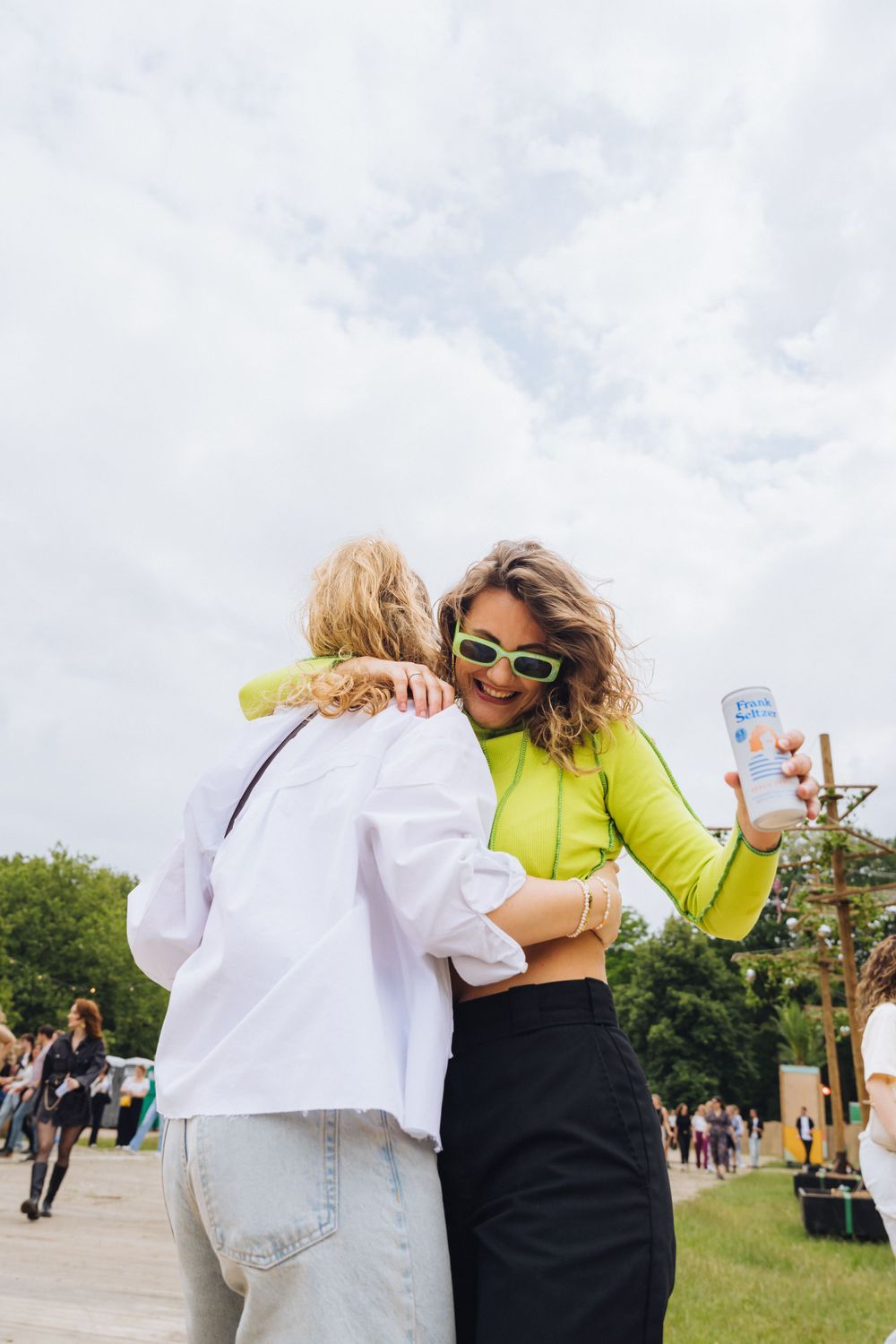 Two friends in casual attire share a joyful moment at an outdoor event while one holds a mobile phone.