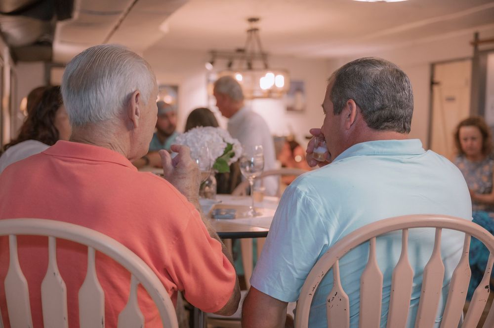 Two people in casual attire sit at a dining table in a restaurant with warm lighting and white chairs.