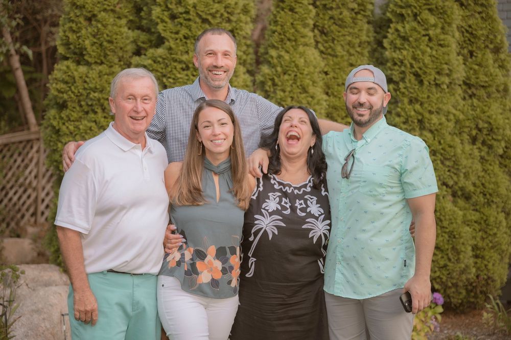 A group of five people pose together outdoors against a backdrop of green shrubs, sharing laughter and smiles.
