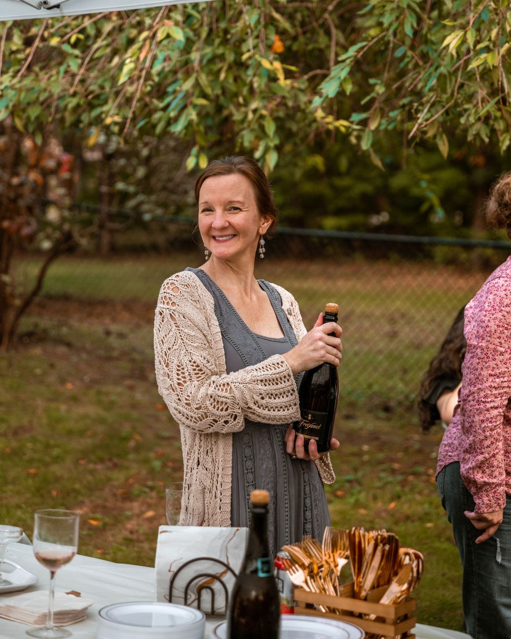 A person holds a wine bottle at an outdoor gathering with autumn trees in the background.