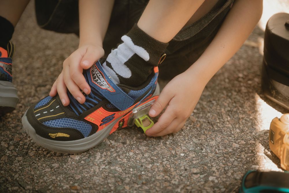 Child putting on colorful blue and orange sneakers with velcro straps while sitting on pavement outdoors.