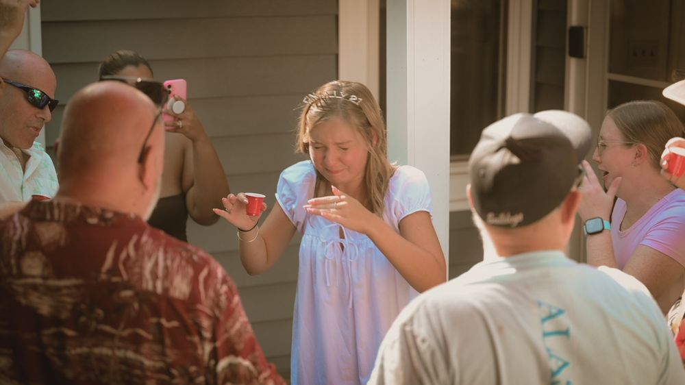 Woman in blue dress holds red cup at outdoor gathering with friends on a porch during golden hour.