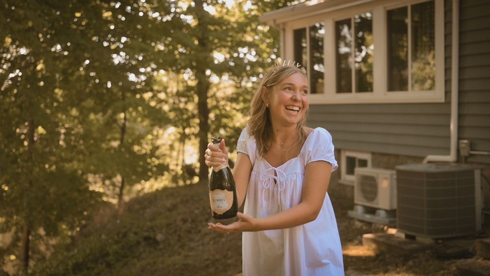 Smiling blonde woman in white dress holding champagne bottle outdoors near trees and house.