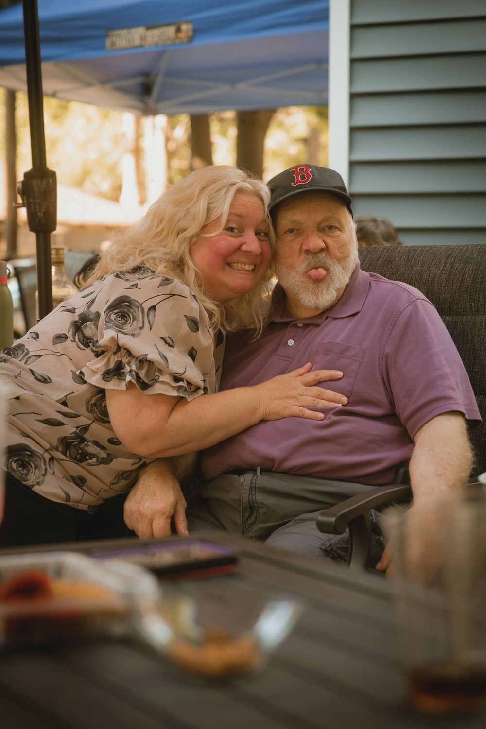 A smiling woman hugs a man in a Boston Red Sox cap making a funny face at an outdoor patio table.