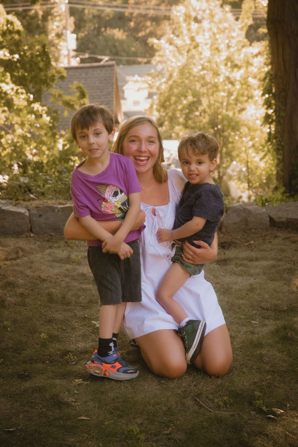 A smiling woman in a white dress kneels on grass hugging two young boys outdoors in a sunny backyard.