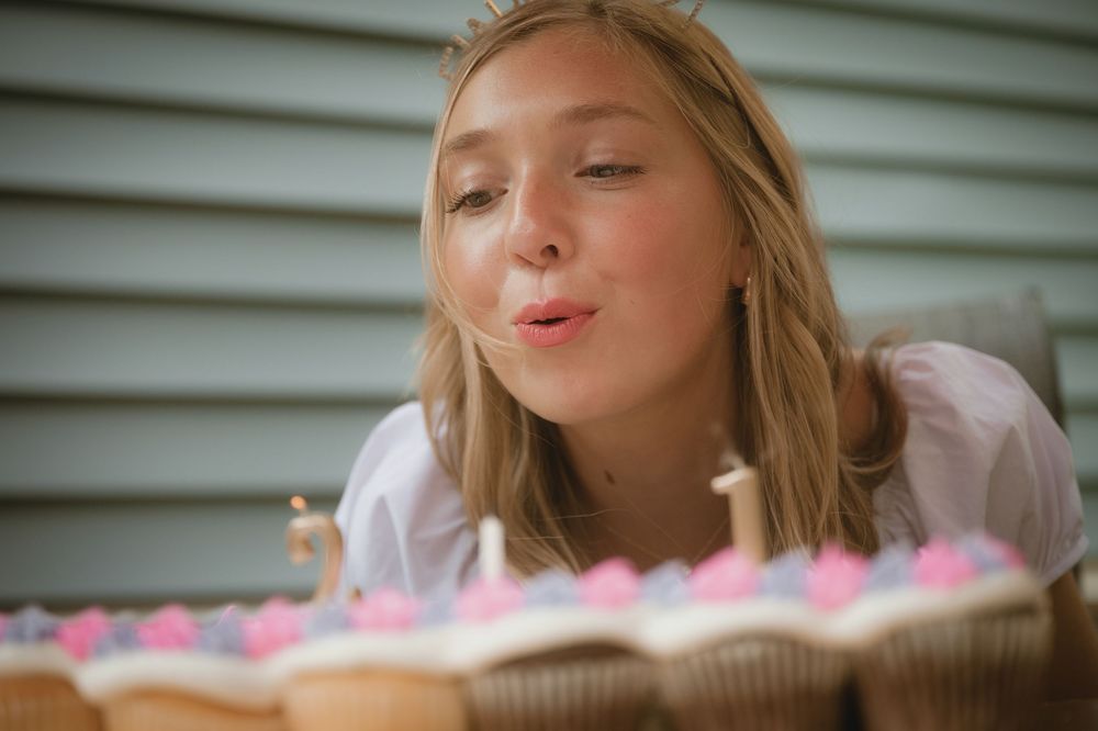 Young blonde girl blowing out birthday candles on decorated cupcakes at an outdoor celebration.