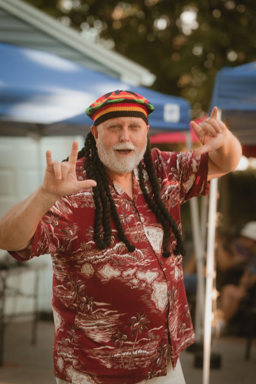 Older man with dreadlocks and Rastafarian hat wearing Hawaiian shirt making shaka signs outdoors.