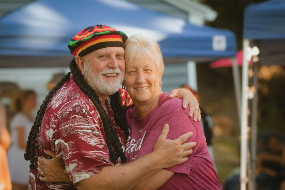 Two smiling seniors embrace warmly at an outdoor event, man wearing Rasta hat and Hawaiian shirt.