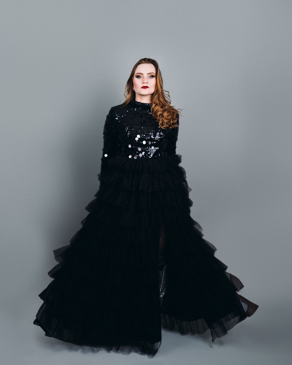 A dramatic fashion portrait of a model wearing a voluminous black tulle gown against a gray studio backdrop.