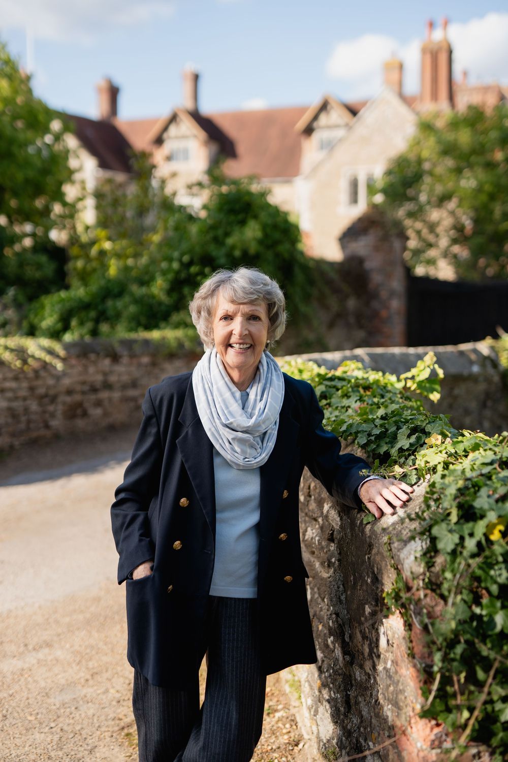 Elderly woman in navy blazer and gray scarf walking along garden path with stone wall and houses.