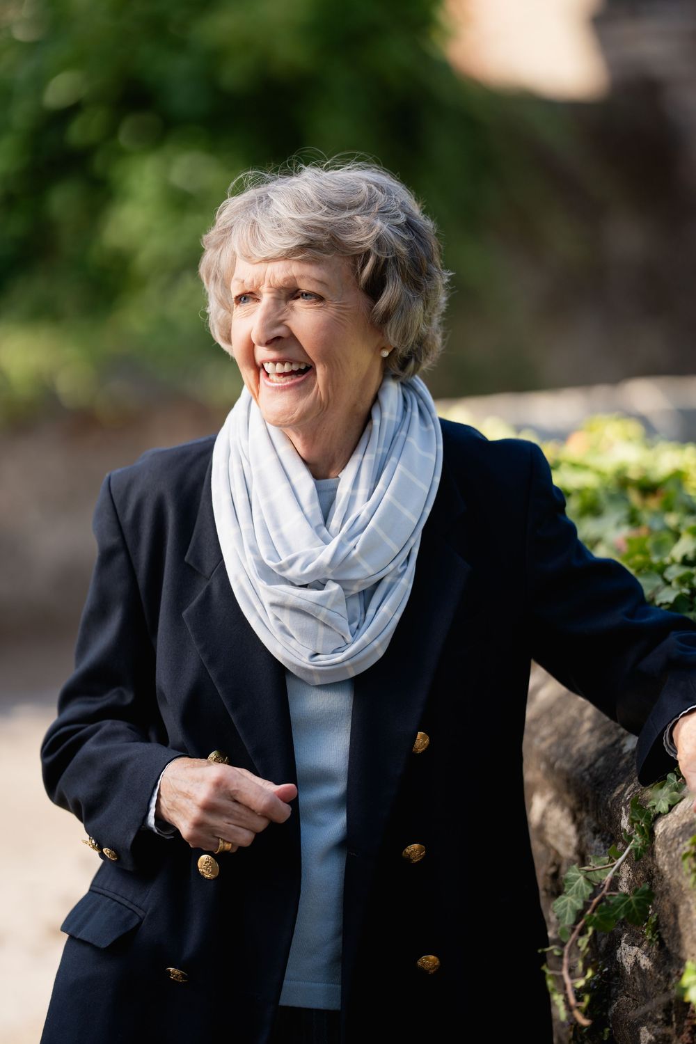 Woman in navy jacket and light scarf smiling outdoors near garden wall.