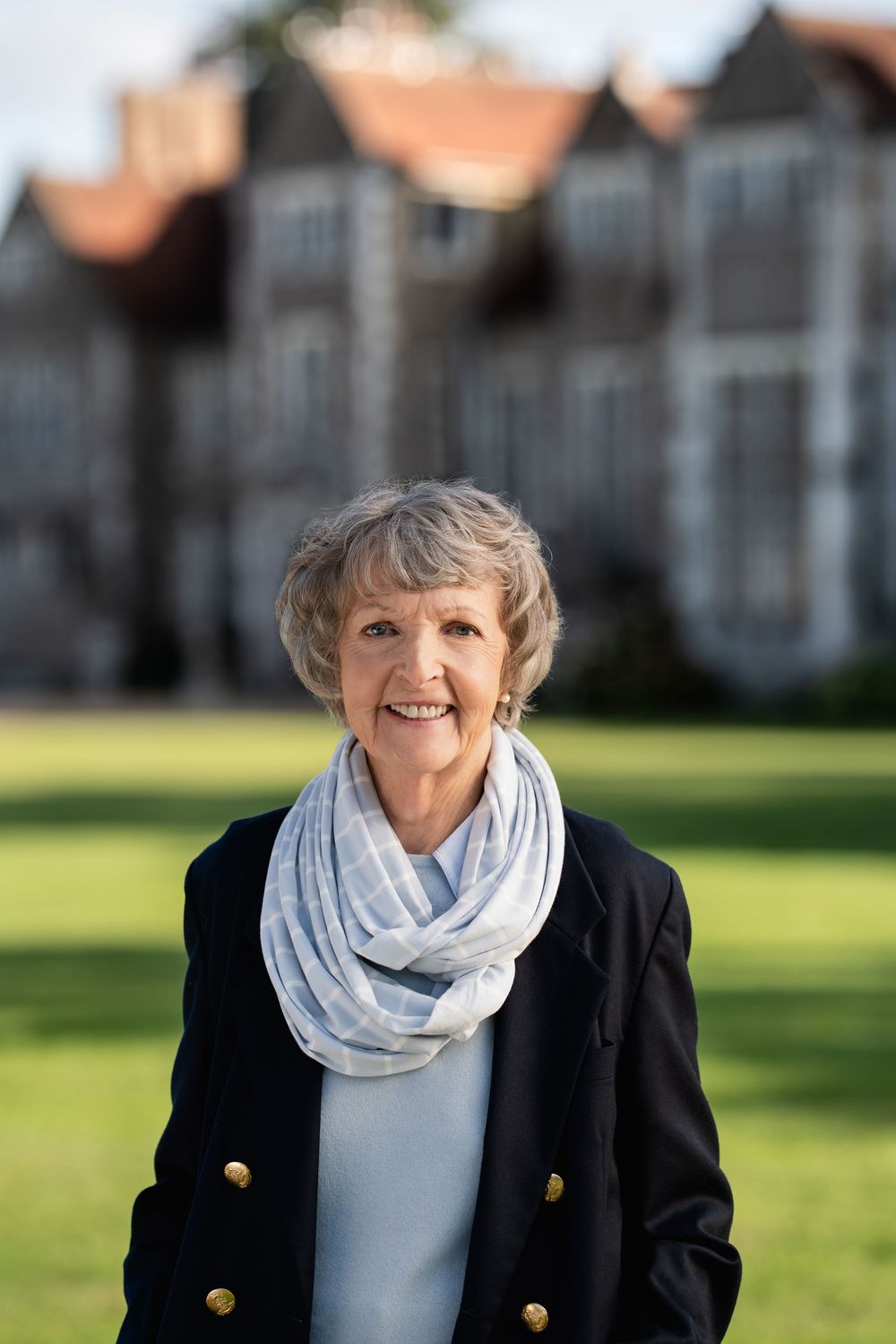 Woman in navy blazer and gray infinity scarf standing outside with buildings in background.