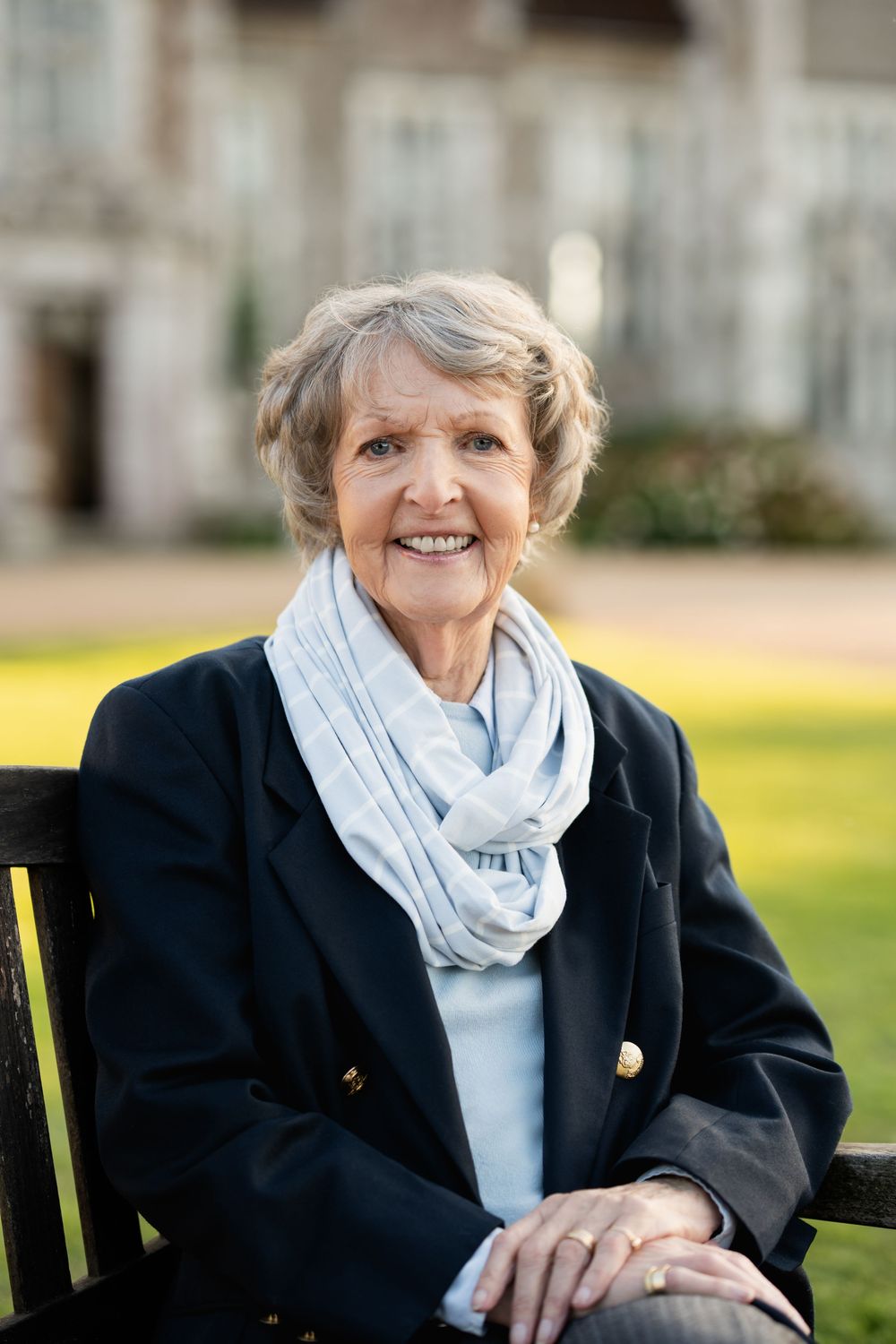 A person in a black blazer and light blue scarf smiles warmly while sitting outdoors on a bench.