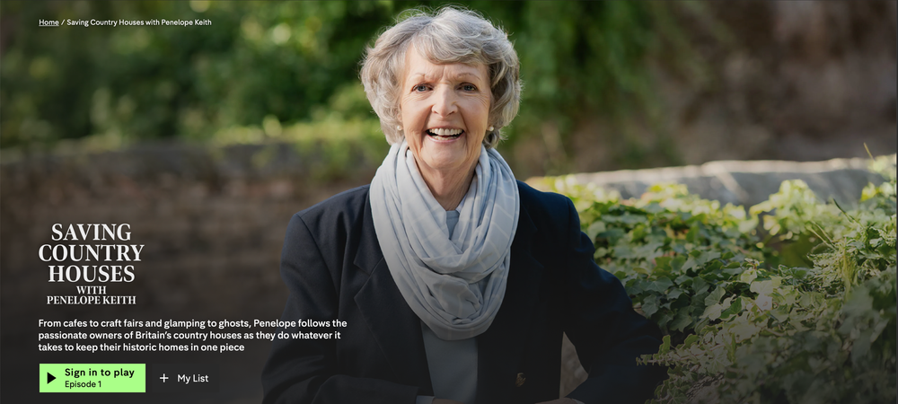 Smiling person in gray scarf and black top stands outdoors near green shrubs in natural lighting.