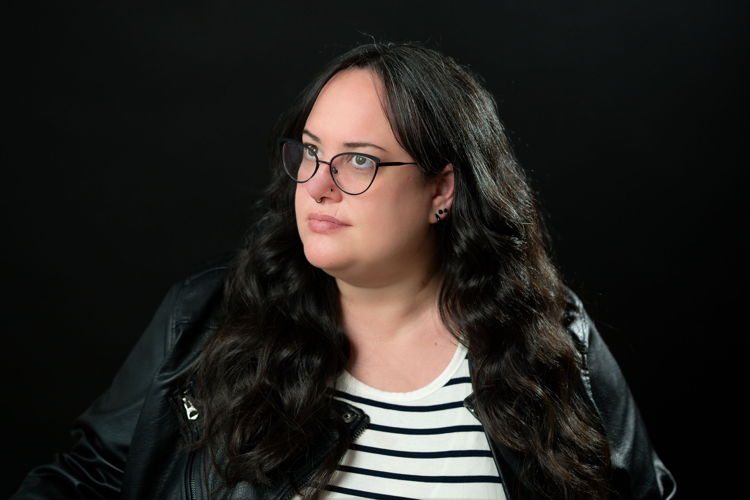Young woman with long dark wavy hair and glasses wearing a striped shirt and black leather jacket.
