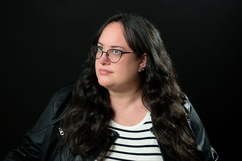 Young woman with long dark wavy hair and glasses wearing a striped shirt and black leather jacket.