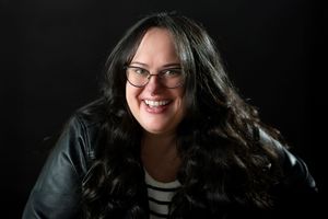 Smiling young woman with long dark wavy hair and glasses in a black leather jacket on dark background.