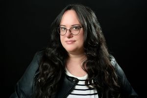 Young woman with long dark curly hair and glasses wearing a black leather jacket and striped shirt.