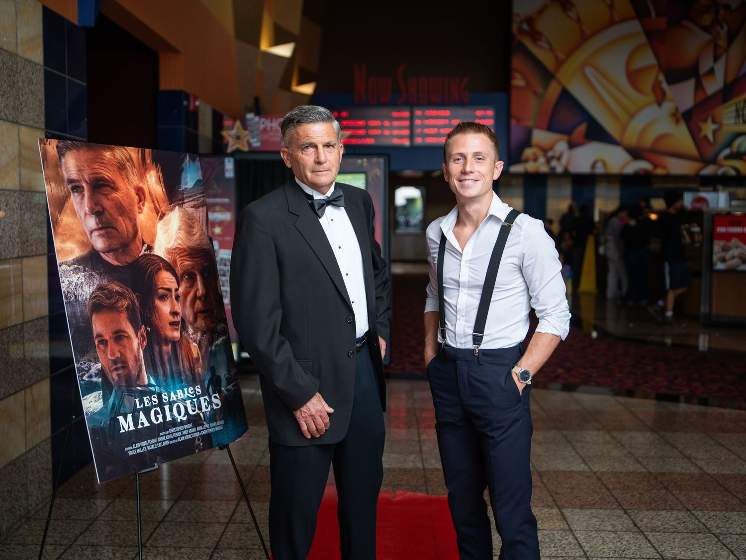 Two men in formal attire stand on a red carpet in front of a movie poster at a theater or cinema event.