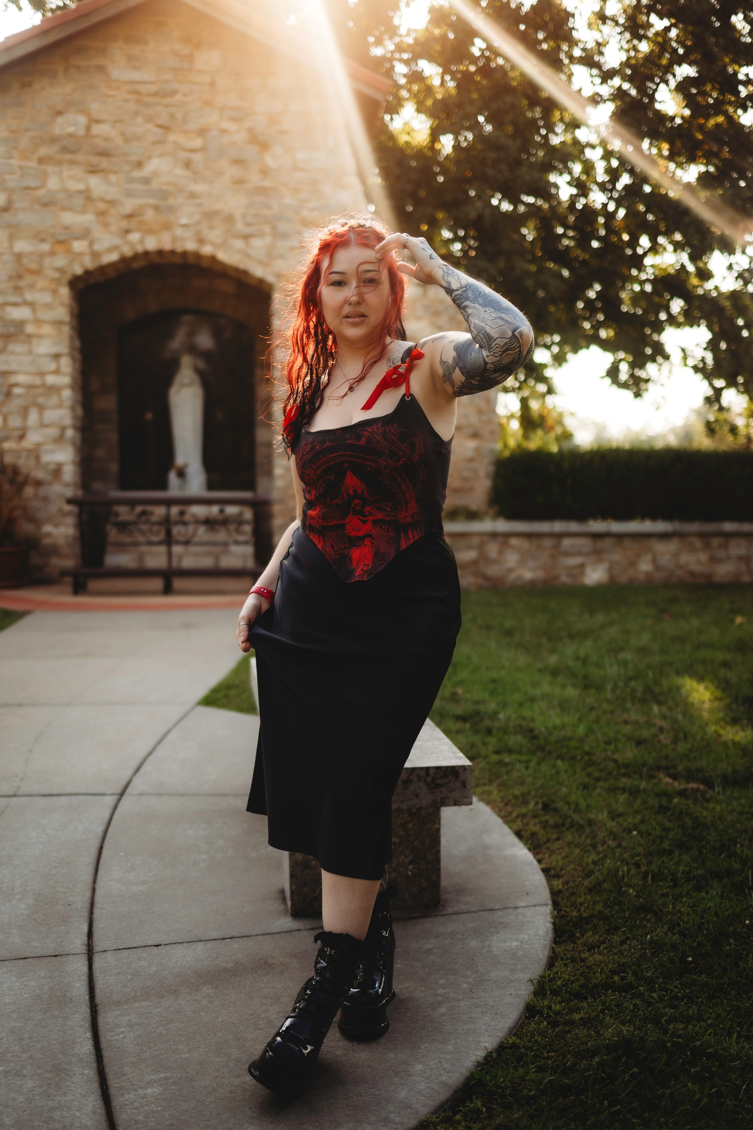 Woman with red hair and tattoos in red corset posing outside stone chapel at sunset. Located in Perryville MO