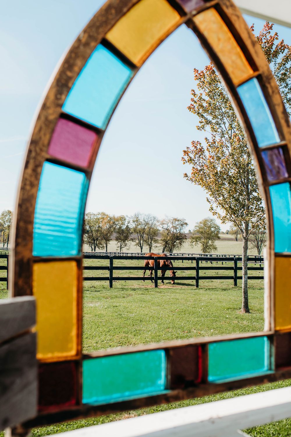 View through a colorful stained glass window frame looking out onto a grassy field with trees in the distance.