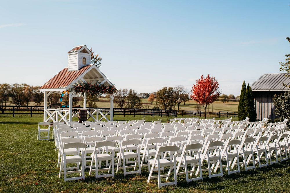 White wedding chairs arranged in rows on a green lawn with a rustic chapel and autumn trees in the background.