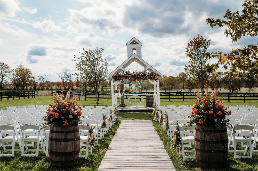 White garden chairs arranged in rows along a wooden walkway with coral floral arrangements and wine barrels.