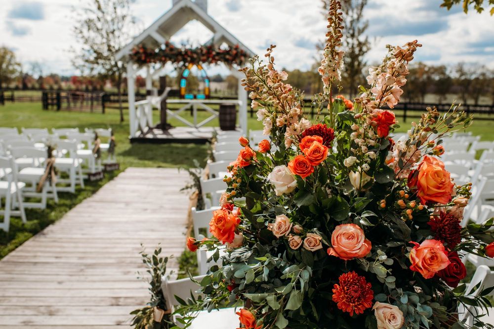 Rustic outdoor wedding venue with white chairs and floral arrangements leading to a white chapel gazebo.