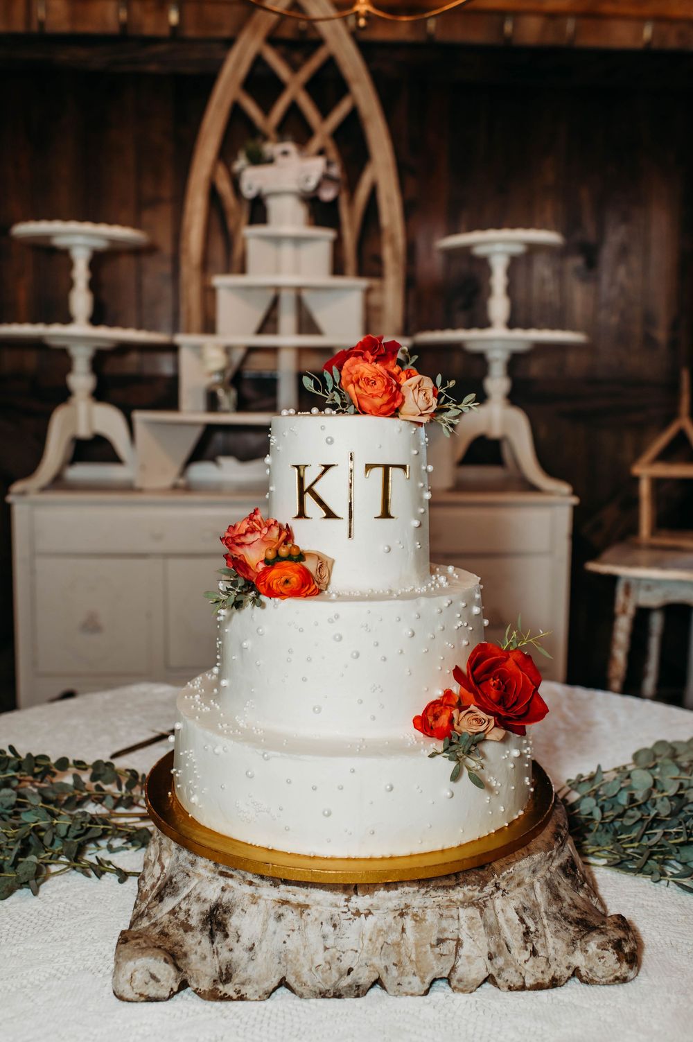 Three-tier white wedding cake with coral roses and 'KT' monogram displayed on rustic wood slice base with vintage decor.