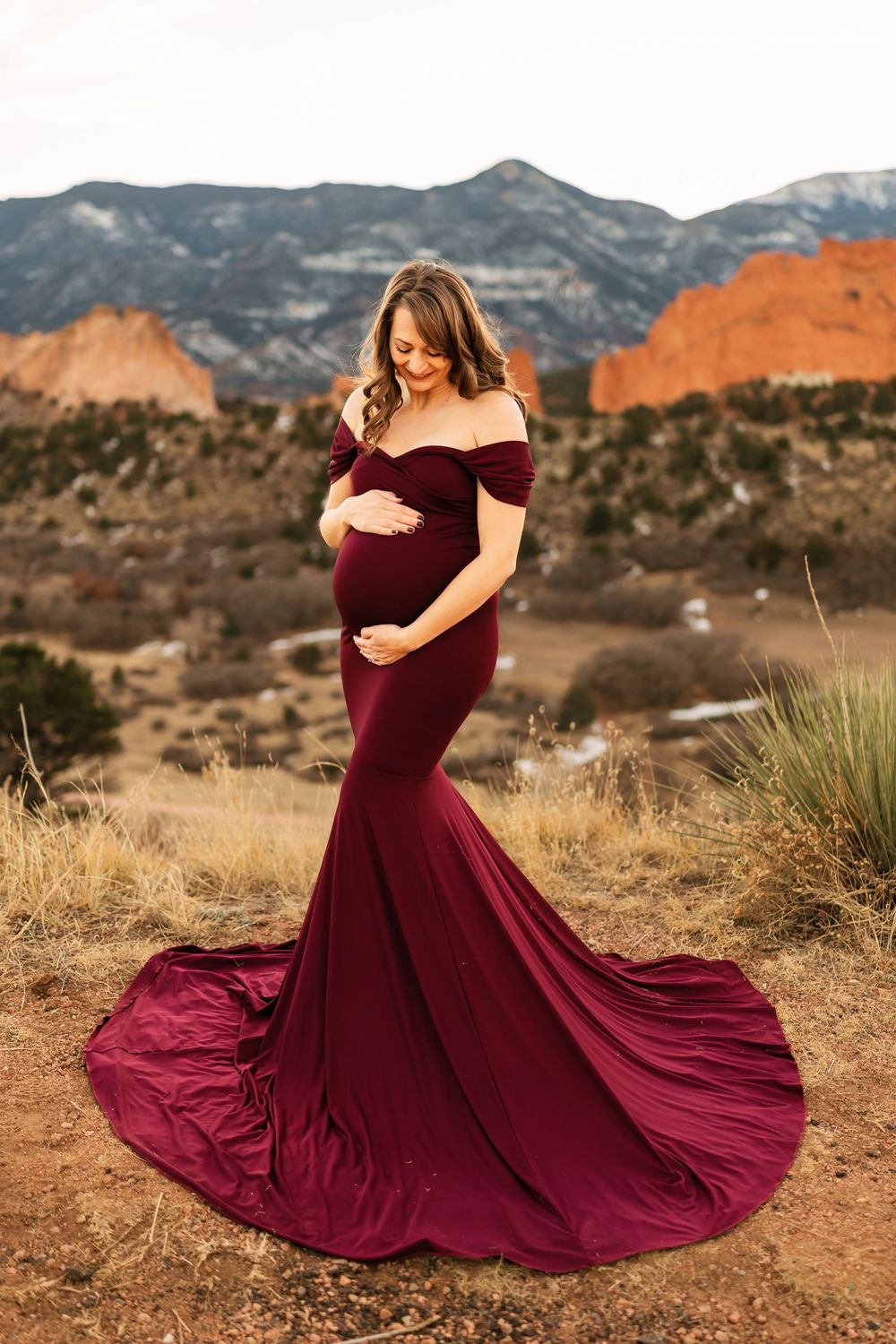 A burgundy formal dress flows dramatically in the desert landscape with mountains in the background.