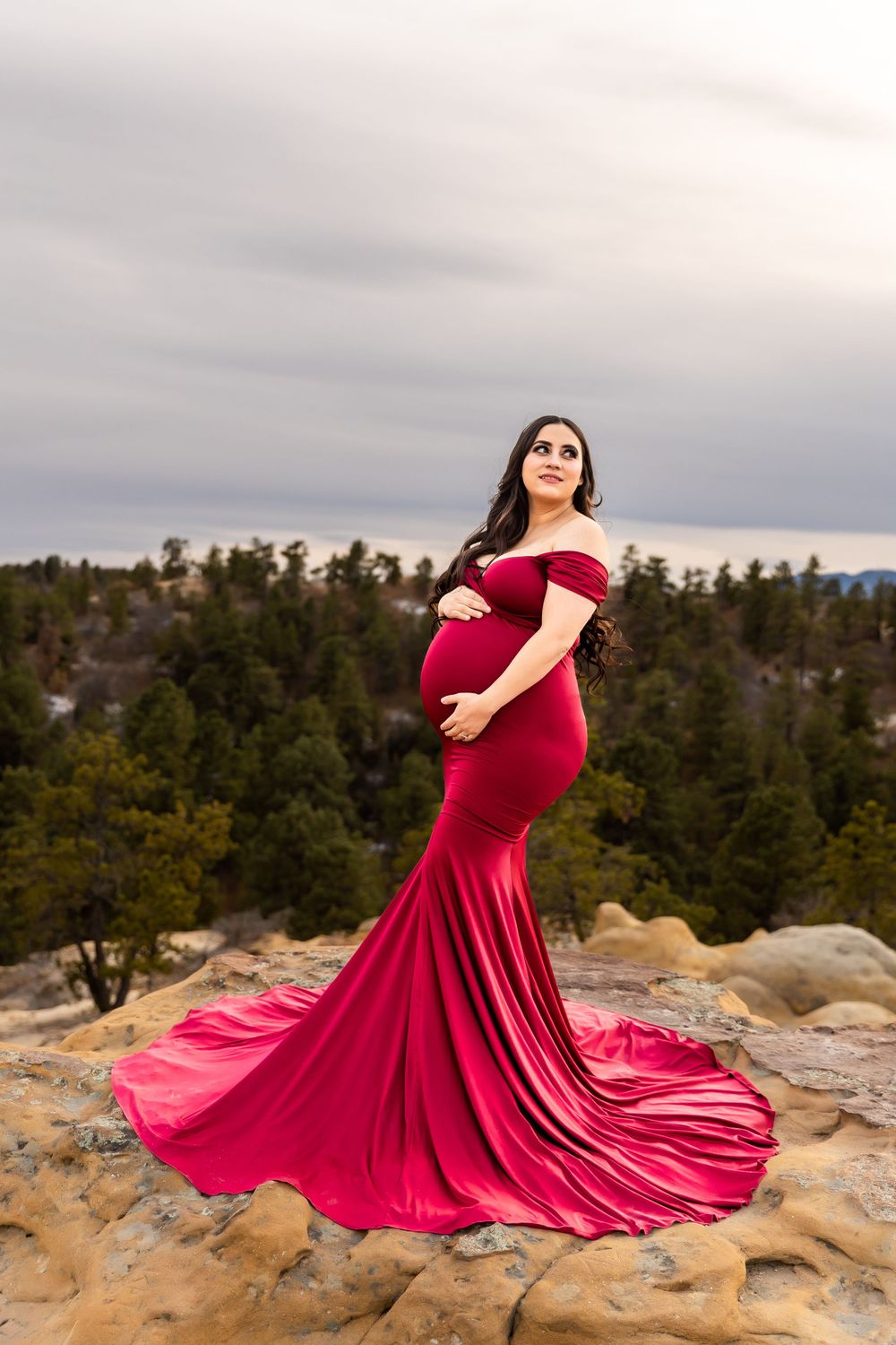 A woman in a flowing red dress twirls gracefully on rocky terrain against a backdrop of evergreen trees and cloudy skies.