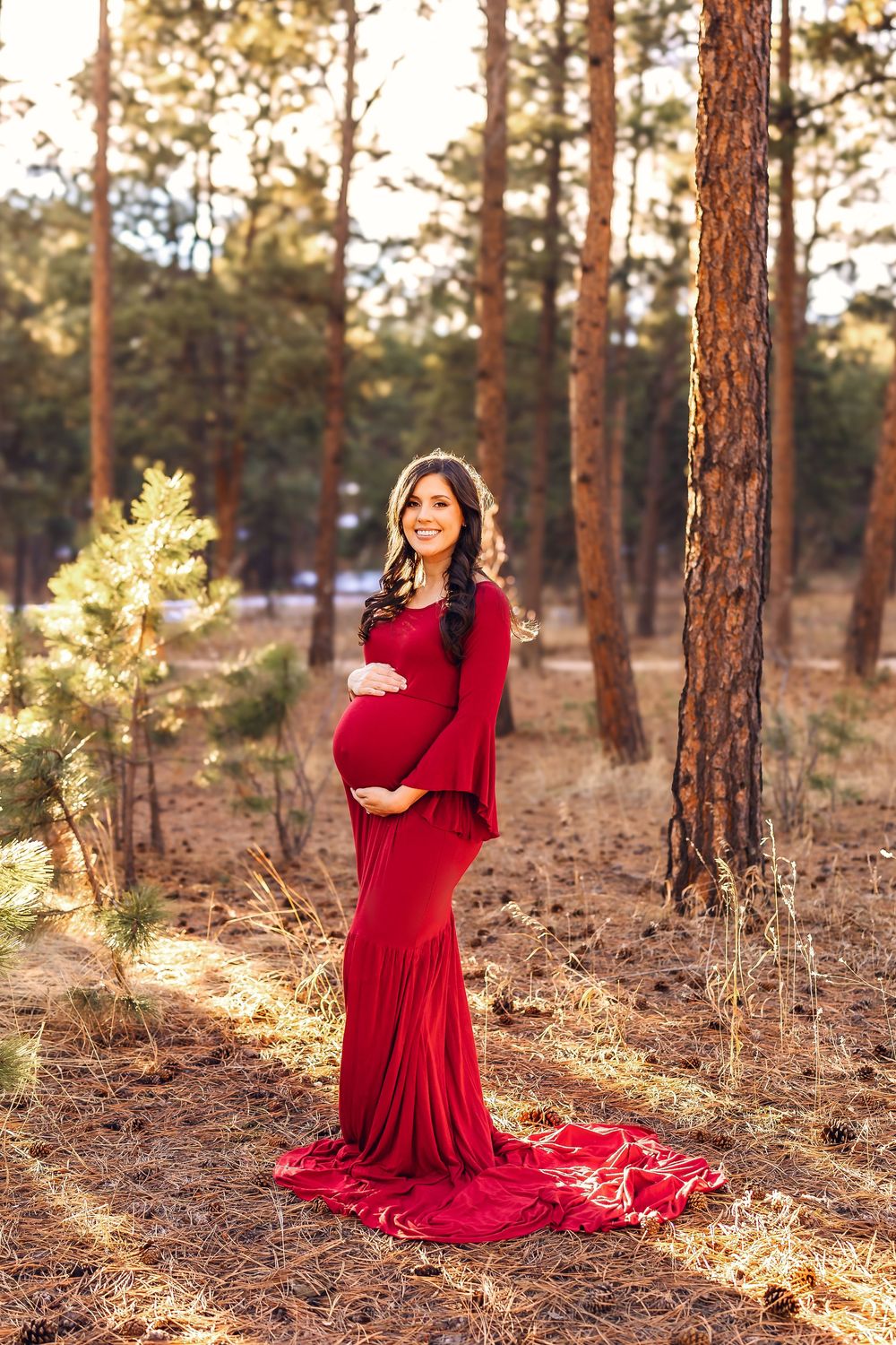 A figure in a flowing red dress poses gracefully among tall pine trees in golden sunlight.
