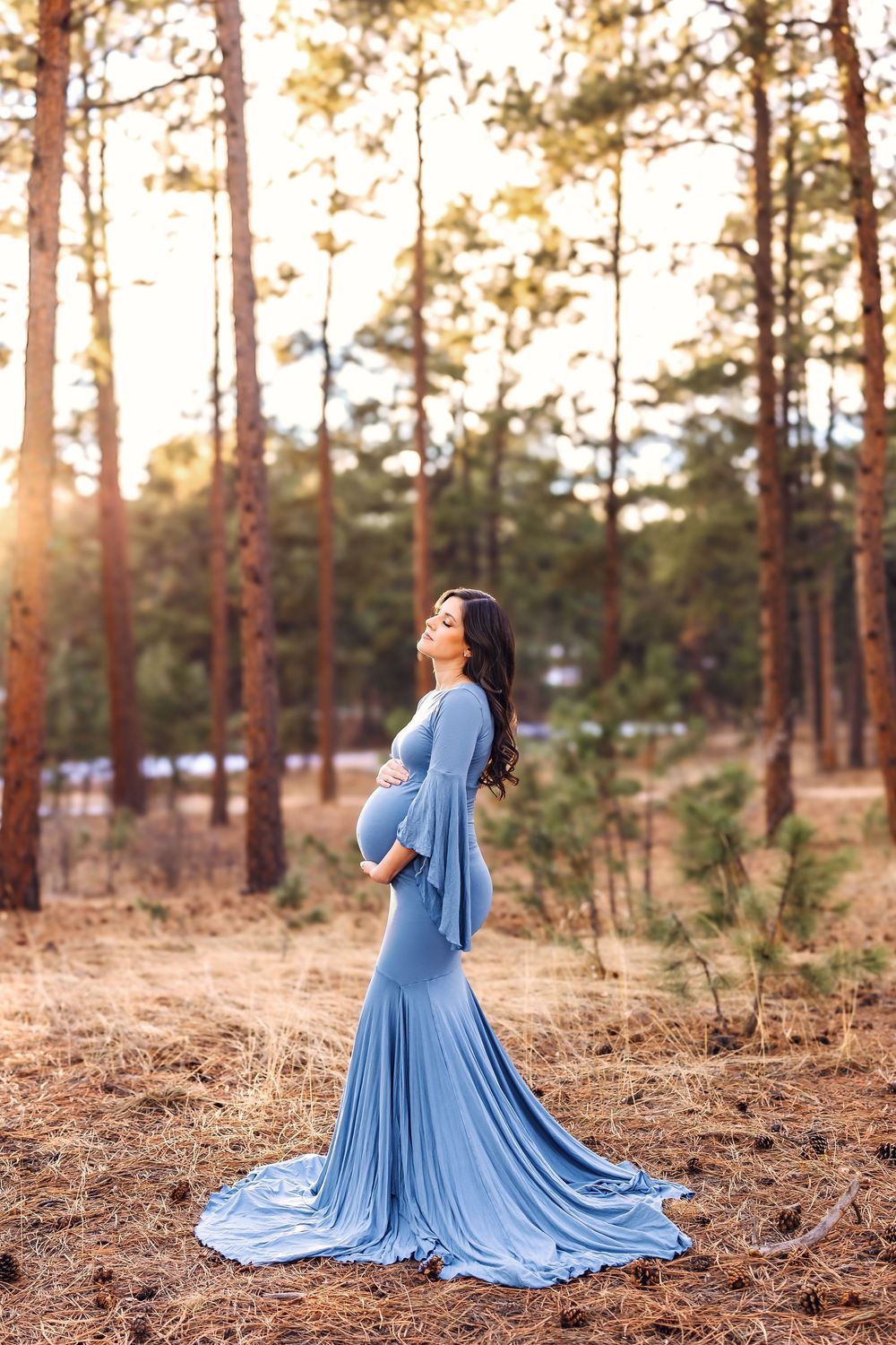 A flowing blue dress creates an ethereal scene against a backdrop of tall pine trees at sunset.