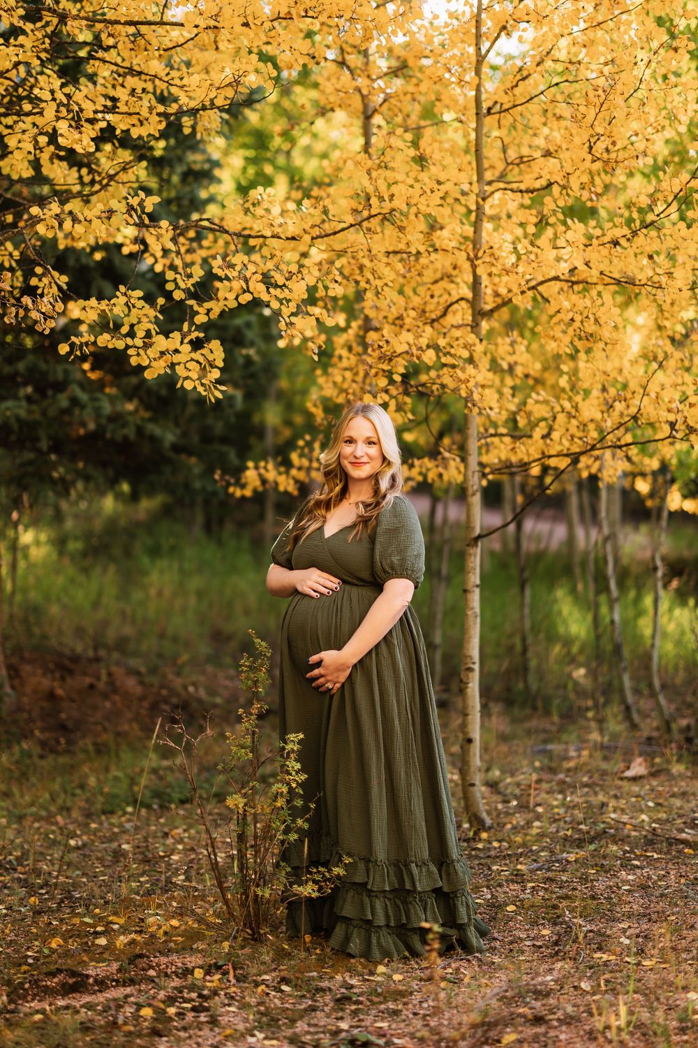 A maternity photo taken in autumn shows an expectant mother in a flowing olive dress against golden fall foliage.