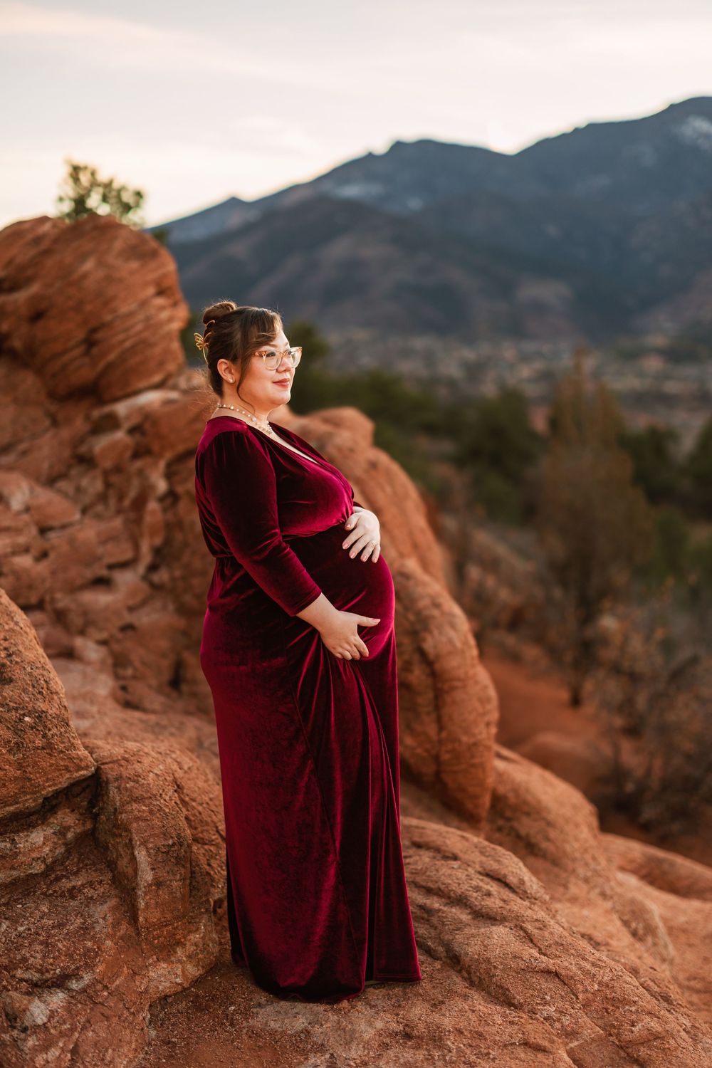 A pregnant woman in a burgundy velvet dress poses on red rocks at sunset with mountains in the background.