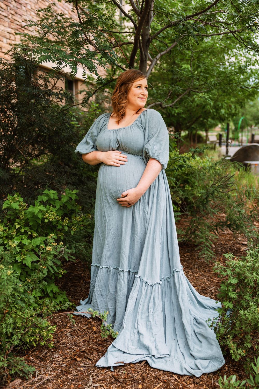 A light blue flowy maxi dress with ruffled tiers is showcased in an outdoor garden setting with green foliage.