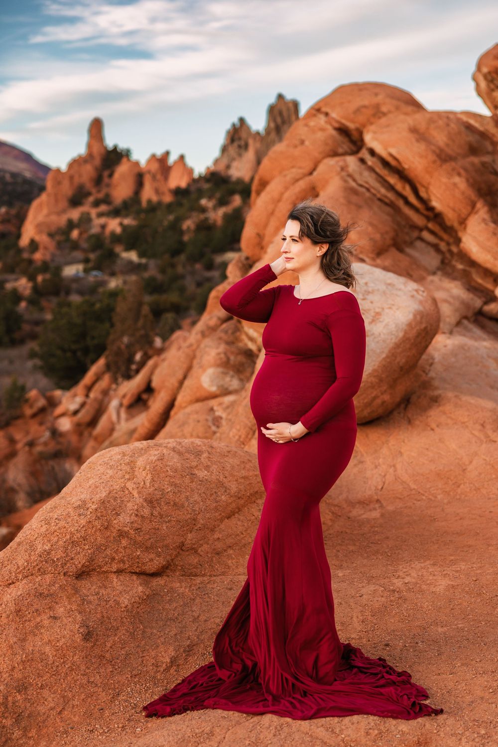 A pregnant woman in a flowing red dress poses against dramatic red rock formations at sunset in Garden of the Gods.