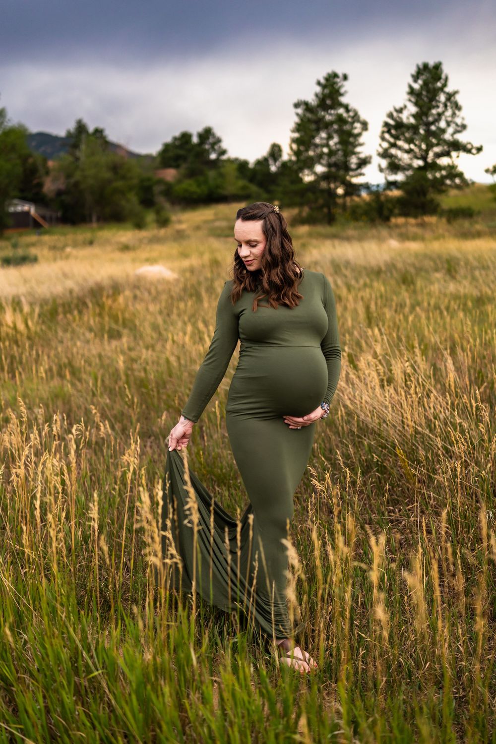 A pregnant woman in a green dress stands thoughtfully in a golden field of tall grass at sunset.