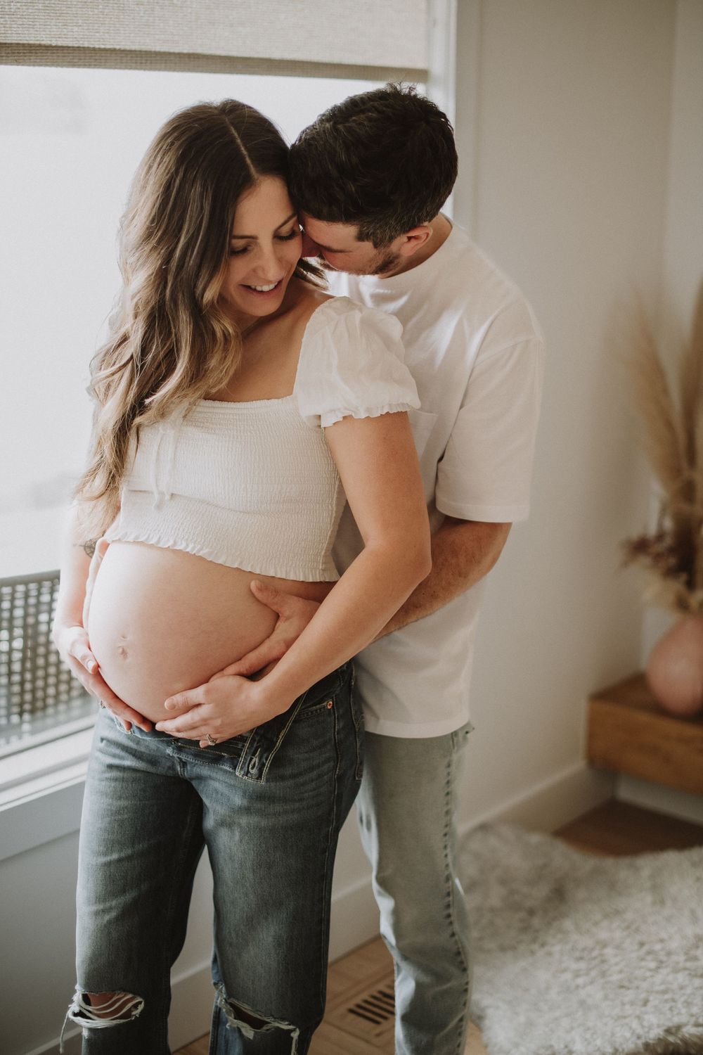 Intimate indoor maternity photo showing expecting parents in casual clothing embracing near a window.